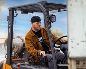 man with a black beanie and brown jacket driving a forklift.